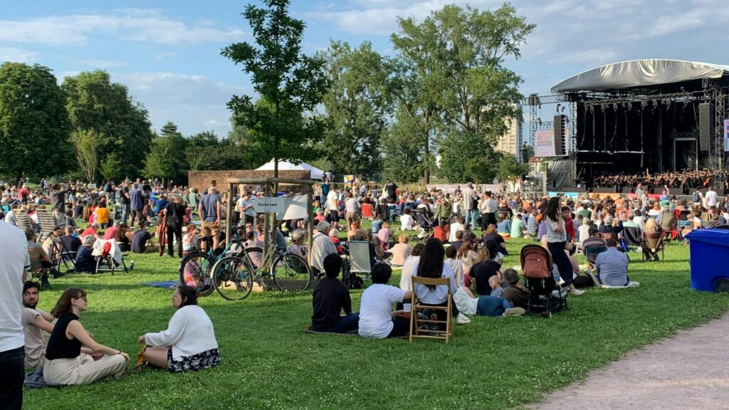 Daytime festival crowd relaxing on a park lawn with an outdoor stage behind the trees