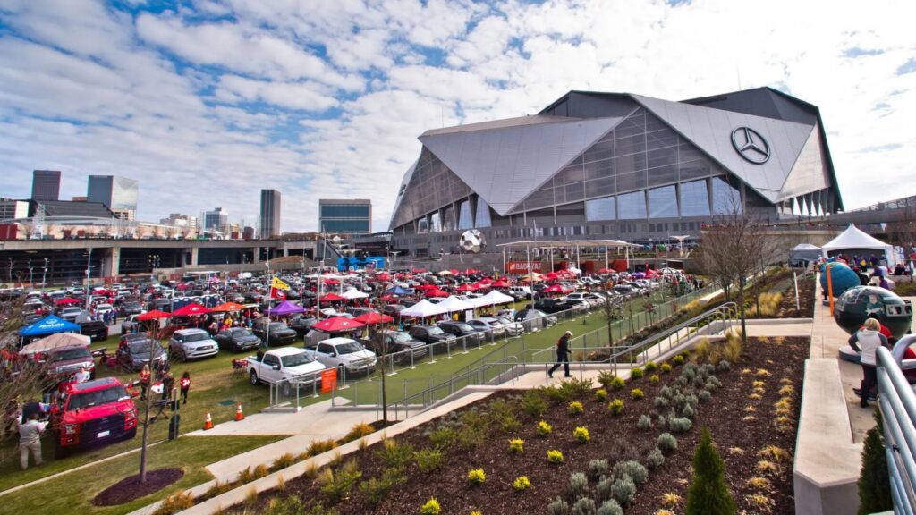 Football fans tailgating on the lawn beside Mercedes-Benz Stadium with the downtown Atlanta skyline behind